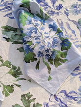 Load image into Gallery viewer, A folded white fabric napkin or tea towel with a watercolor-style botanical print in blue and green, laid on top of a larger matching textile with a cream base and blue floral pattern.
