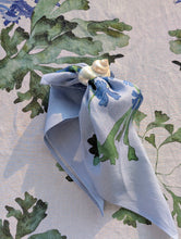 Load image into Gallery viewer, A light blue linen napkin with a green and blue floral print, tied with a small seashell on a patterned tablecloth.
